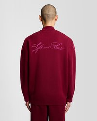 Maglione oversized color bordeaux con colletto e polsini a costine, caratterizzato da ricamo rosa "Style and Scott" sulla parte posteriore. Texture liscia.
