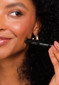 Musta meikkikynä pidetään poskea vasten, mallilla kiharat hiukset ja kultaiset renkorenkaat korvissa. Hienovarainen huuliväri ja luonnollinen meikkipinta.