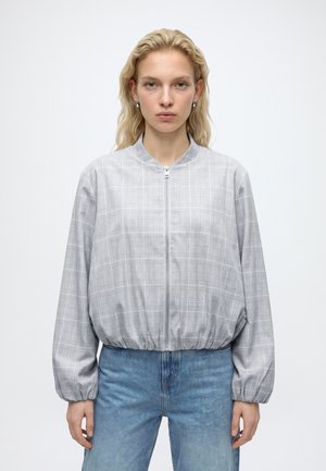 Frau mit welligem blondem Haar trägt eine hellgraue karierte Zip-up-Jacke mit elastischen Bündchen und blaue Jeans, stehend vor einem schlichten weißen Hintergrund.