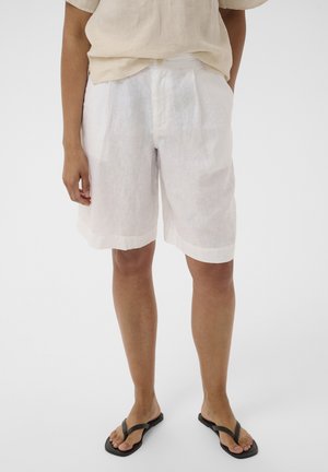 Person iført løse hvide shorts til knæet, beige kortærmet top og sorte flip-flops, stående foran en ensfarvet baggrund.