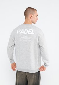 Grå sweatshirt med lange ærmer, der har hvid tekst på ryggen: "PADEL CROWN CHAMPIONSHIP." Ribbet manchet og kant. Casual pasform.