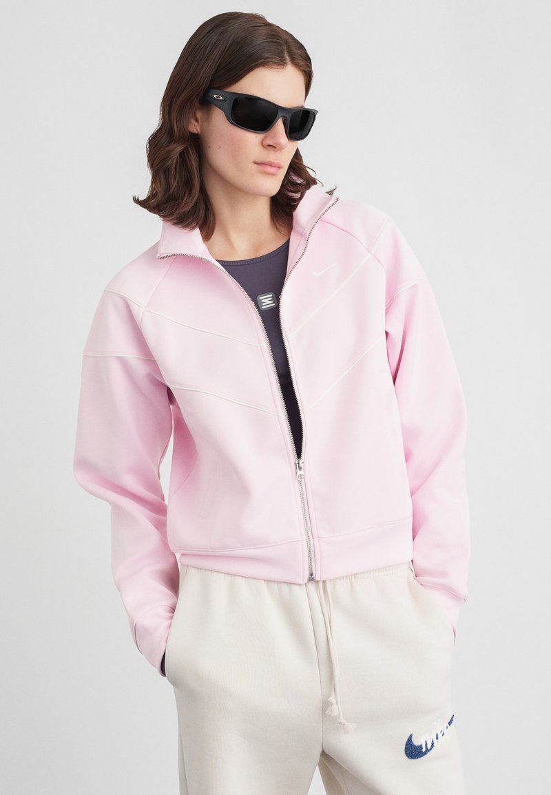 Άτομο που φοράει μαύρα γυαλιά ηλίου, ανοιχτό ροζ φερμουάρ μπουφάν, σκούρο γκρι μπλούζα και κρέμα παντελόνια φόρμας με λογότυπο Nike, χέρια στις τσέπες.