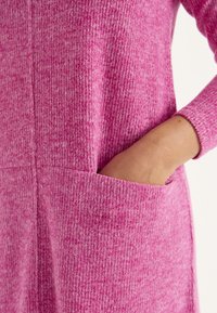 Rosa gestricktes Kleid mit vertikaler Rippstruktur, ausgestattet mit seitlichen Taschen und taillierten Ärmeln. Das Material wirkt weich und leicht strukturiert.