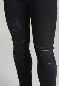 Jeans negros ajustados con desgastes deshilachados en las rodillas y el muslo, fabricados en denim con una textura suave y un diseño ajustado.