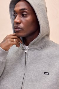 Sweat à capuche gris zippé avec capuche, fabriqué en tissu doux en mélange de coton, présentant une petite étiquette logo noire sur la poitrine et des poignets côtelés.