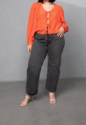 Femme portant un chemisier orange à manches longues avec des pois blancs, un jean noir taille haute et des talons beiges ouverts devant sur fond gris.