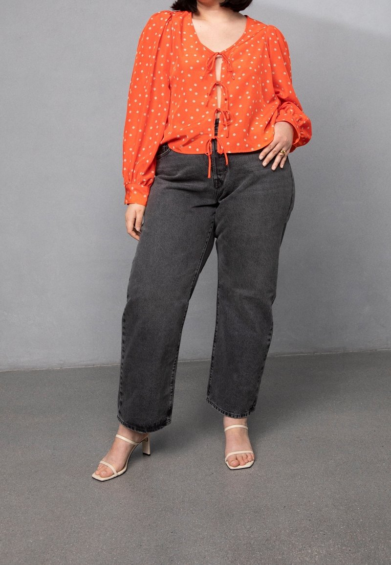 Femme portant un chemisier orange à manches longues avec des pois blancs, un jean noir taille haute et des talons beiges ouverts devant sur fond gris.