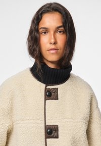 Jauna sieviete ar viļņainiem tumšiem matiem, valkājot krēmkrāsas šerpas jaku ar melniem pogām un melnu ribotu augstu apkakli.