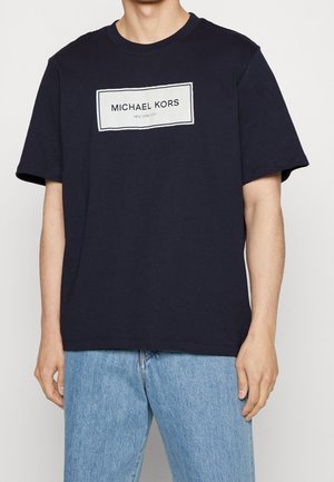 Man in een marineblauwe Michael Kors T-shirt met een wit rechthoekig logo en lichtblauwe spijkerbroek, staand tegen een effen achtergrond.