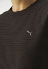 Schwarzes geripptes T-Shirt mit Rundhalsausschnitt. Hat ein kleines graues gesticktes Logo auf der Brust. Der Stoff wirkt weich und leicht strukturiert.