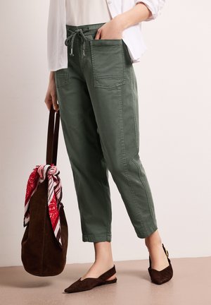 Osoba nosząca przycięte oliwkowozielone spodnie z przednimi kieszeniami i brązowe baleriny na pasku, trzymająca brązową zamszową torbę z czerwoną i białą bandaną.