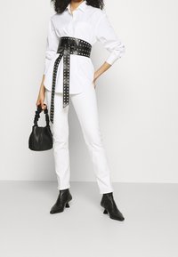 Camicia bianca con bottoni abbinata a pantaloni bianchi a vita alta, cintura in pelle nera con borchie e borsa nera con manico intrecciato; stivali neri alla caviglia.