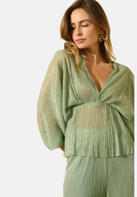 Blouse plissée vert clair avec un décolleté en V profond et des manches chauve-souris, présentant une texture scintillante et un design lâche et fluide.