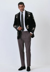 Black velvet blazer with floral embroidery, white shirt, patterned tie, grey trousers, and black shoes. Formal, tailored fit.