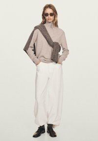 Vrouw in zonnenbril, beige coltrui, gebreide sjaal over schouder, witte wijde broek en zwarte nette schoenen.