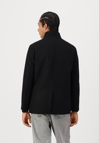 Svart ullkåpe med høy krage, rett snitt og subtilt bakslipp, sett fra baksiden. Kombinert med grå jeans.