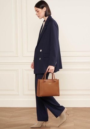 Marineblauwe dubbelgestikte blazer, bijpassende broek, beige haklaarzen tot de enkels en een gestructureerde bruine handtas met bovenhandvatten.