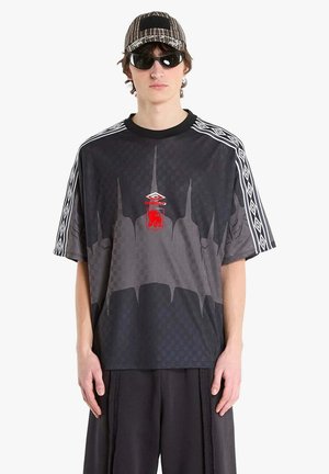 Giovane uomo che indossa una t-shirt oversize con motivo geometrico nero e grigio, pantaloni larghi abbinati, cappello a righe e occhiali da sole neri, in piedi davanti a uno sfondo bianco.