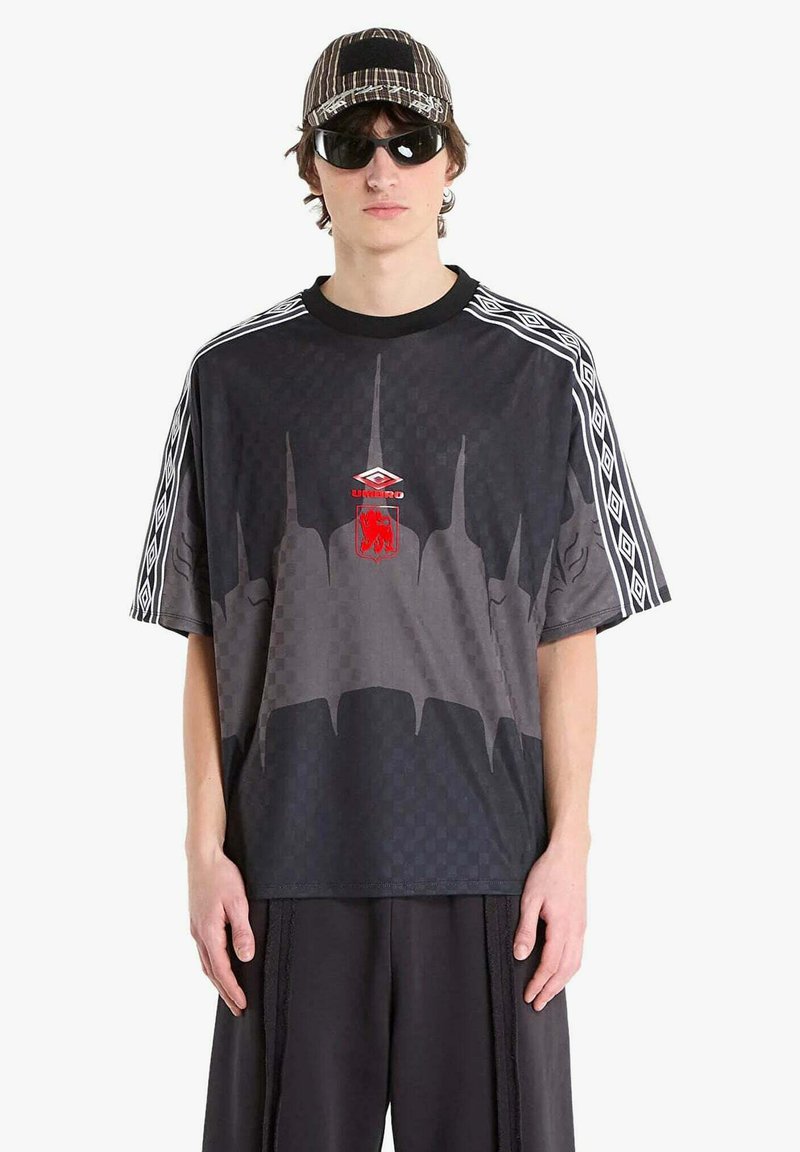 Giovane uomo che indossa una t-shirt oversize con motivo geometrico nero e grigio, pantaloni larghi abbinati, cappello a righe e occhiali da sole neri, in piedi davanti a uno sfondo bianco.