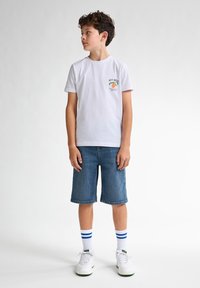 Vit bomulls t-shirt med "KEY WEST" grafisk tryck, blå denimshorts, vita sneakers och randiga strumpor. Stående position mot en ljus bakgrund.