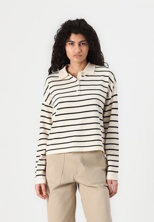 Polo a maniche lunghe a righe crema e nere con colletto e due bottoni, abbinato a pantaloni beige con tasche a toppa.