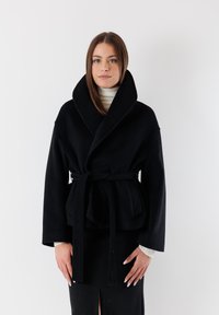 Cappotto nero con cintura in vita, colletto sciallato, maniche lunghe e legatura in vita. Texture liscia, design minimalista e forma strutturata.