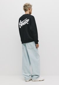 Sort sweatshirt med hvidt skriftlogo på ryggen, parret med lyseblå oversized denimbukser og hvide sneakers.