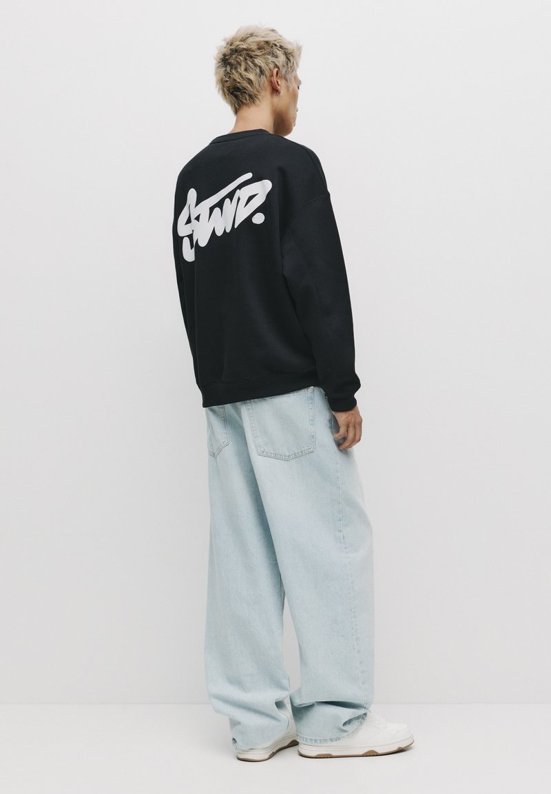 Sort sweatshirt med hvidt skriftlogo på ryggen, parret med lyseblå oversized denimbukser og hvide sneakers.