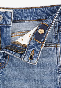 Cintura de vaqueros de mezclilla con costuras azul oscuro, botón de cobre y cremallera. El bolsillo interior muestra una tela blanca con formas impresas.