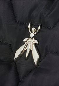 Tessuto trapuntato nero con cuciture geometriche, caratterizzato da un'applicazione a forma di insetto in metallo lucido nei toni argento e oro.