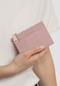 Roze leren portemonnee met een gerimpelde afwerking en gouden hardware, voorzien van een ritssluiting en meerdere kaartslots. Vastgehouden in een hand tegen een lichte achtergrond.