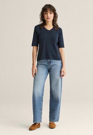 Nainen seisoo päällään tummansininen V-kaula-aukollinen paita, vaaleansiniset suoralahkeiset farkut ja ruskeat loafersit yksivärisen beigen taustan edessä.