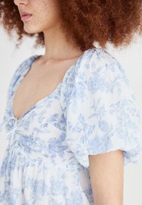 Vestido floral azul claro feito de tecido leve, apresentando mangas bufantes, um decote em V e um detalhe de laço no busto.