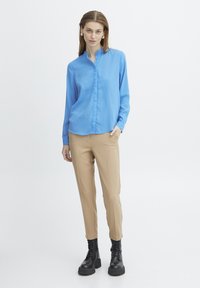 Blusa azzurro chiaro a maniche lunghe con collo alla mandarino, abbinata a pantaloni beige su misura e stivaletti neri con lacci alla caviglia. Finish in tessuto liscio.