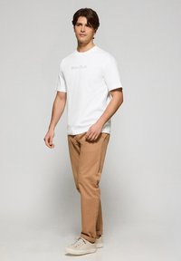 Biała koszulka z krótkim rękawem z logo "Polo Club", w połączeniu z jasnobrązowymi spodniami i beżowymi trampkami, stojąca na neutralnym tle.