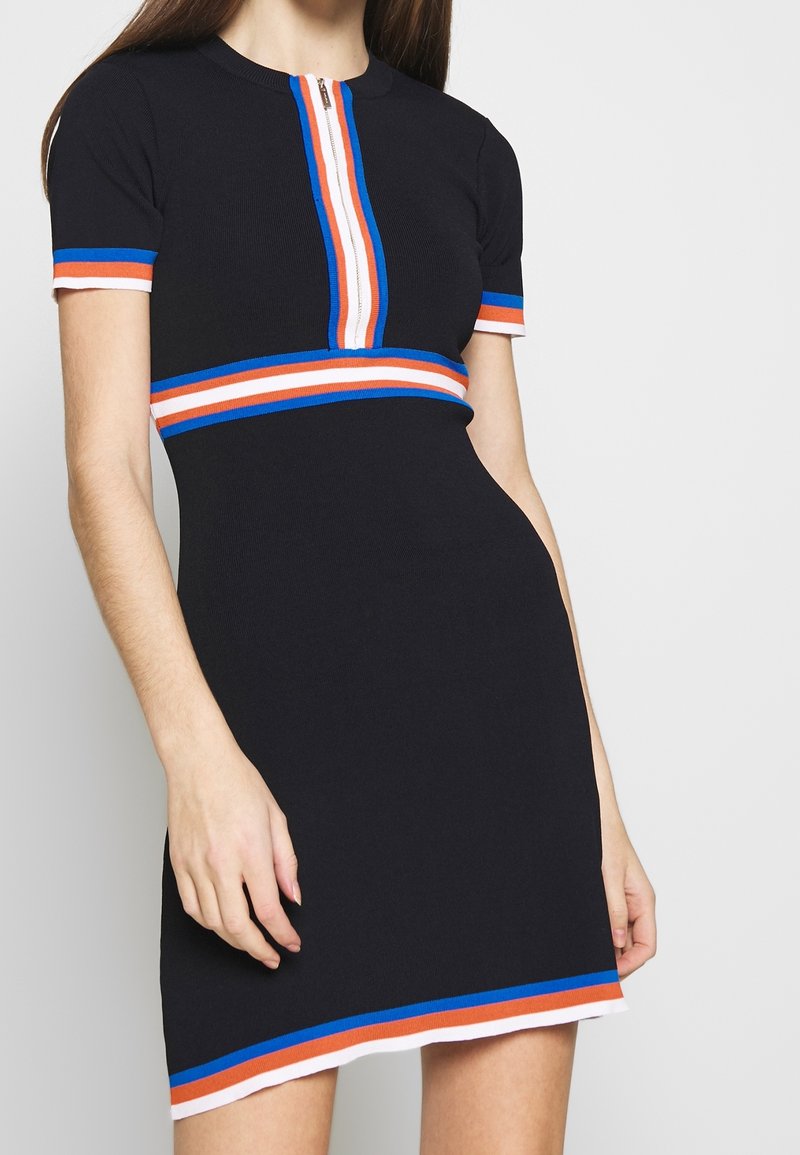 Robe noire à manches courtes avec fermeture éclair sur le devant et rayures bleues, blanches et orange sur les manches, la poitrine, la taille et l’ourlet, portée par une femme.