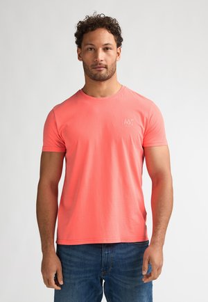 T-shirt en coton corail à manches courtes et col rond. Présente un petit logo blanc sur la poitrine. Associé à un jean en denim bleu.