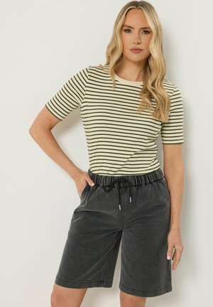 Kvinde med langt blondt hår iført en kortærmet top i grønne og hvide striber og sorte shorts med snøre til knæene.