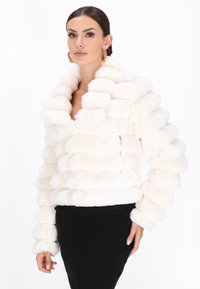 Kvinde iført en hvid, struktureret fake fur-jakke og sort nederdel, med hår trukket tilbage og gyldne hoops øreringe, stående mod en hvid baggrund.