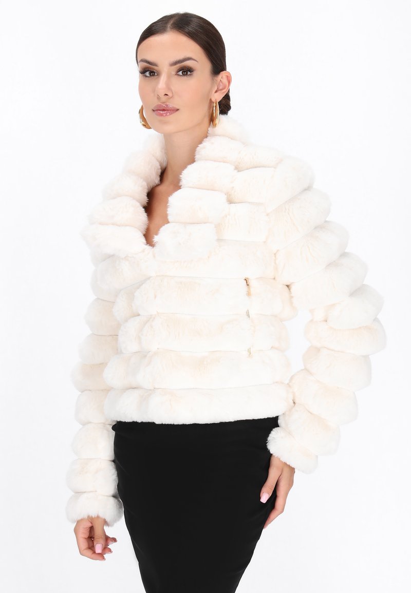Kvinde iført en hvid, struktureret fake fur-jakke og sort nederdel, med hår trukket tilbage og gyldne hoops øreringe, stående mod en hvid baggrund.