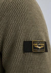 Olivgrüner gestrickter Pullover mit einem strukturierten Muster, versehen mit einem schwarzen rechteckigen Etikett mit einem gelben Logo und Text an der Seite.