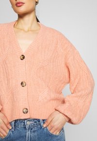 Rosa kabelstickad cardigan med djup V-ringning, prydd med stora runda knappar och ribbade manschetter, bäddad över blå denimjeans.