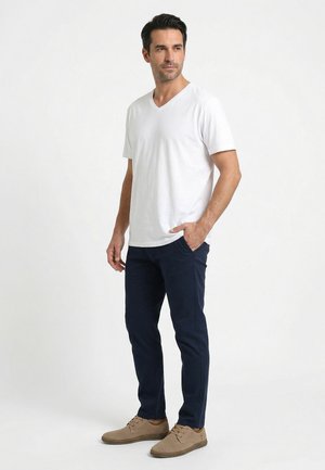 Muž stojící s rukou v kapse, oblečený v bílém tričku s krátkým rukávem a výstřihem do V, tmavě modrých kalhotách a světlých perforovaných společenských botách.