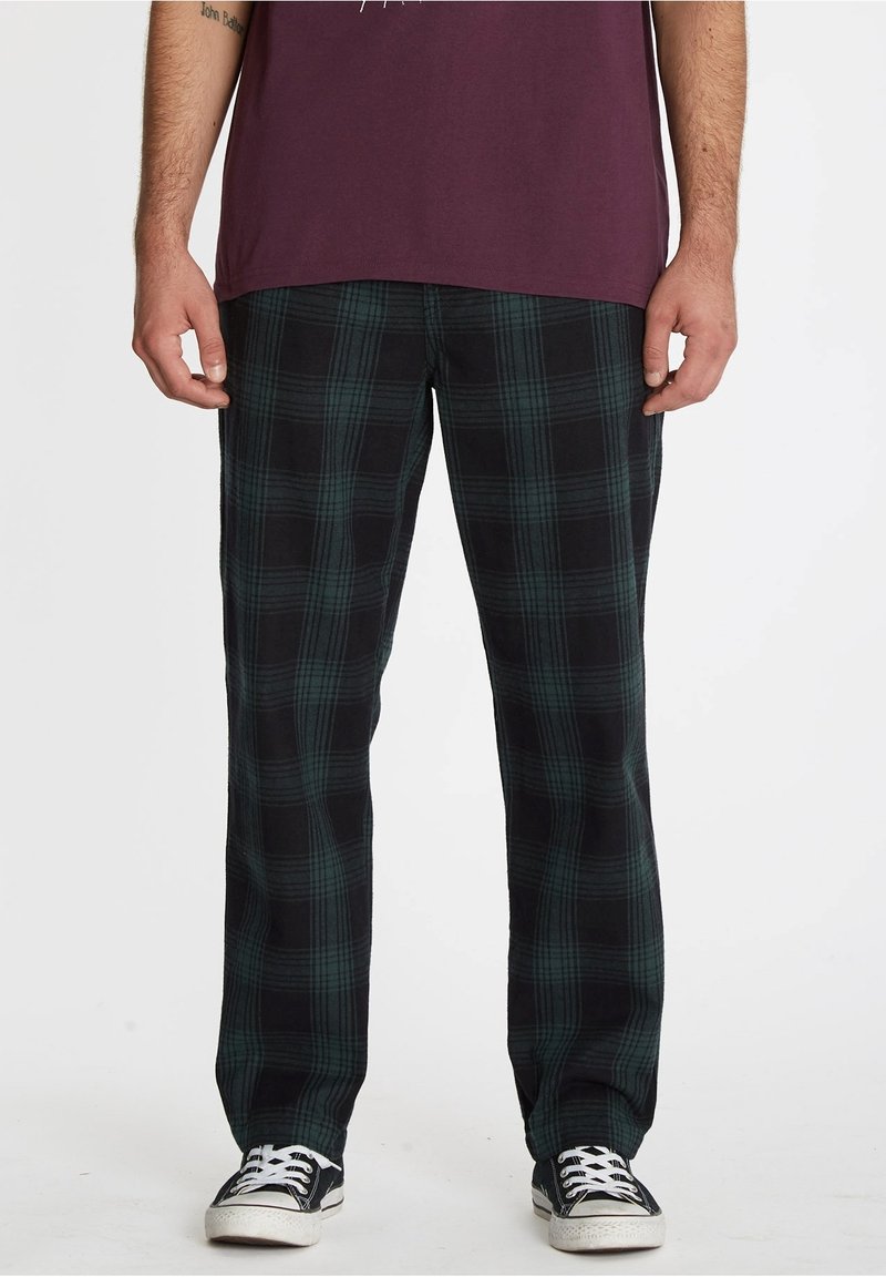 Pantalon à carreaux noir et vert, coupe décontractée, en tissu doux ; porté avec une chemise bordeaux et des baskets noires.