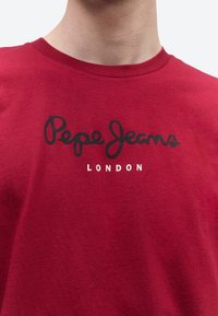 Άνδρας φοράει κόκκινο t-shirt με στρογγυλή λαιμόκοψη, με τη φράση "Pepe Jeans" σε μαύρα γράμματα γραφής και τη λέξη "LONDON" σε λευκά γράμματα μπλοκ στο στήθος.