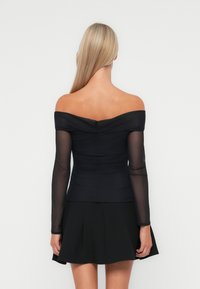 Haut noir dénudé des épaules avec des manches longs transparentes, texture froncée au niveau du buste, et silhouette ajustée. Associé à une jupe noire évasée.