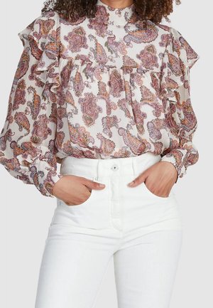 Femme portant un chemisier à manches longues avec volants et motif cachemire violet et orange, rentré dans un pantalon blanc taille haute, mains dans les poches.