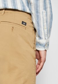 Pantalon chino beige avec une poche latérale, orné d'un petit patch logo noir et une texture coton lisse, assorti à une chemise rayée bleue et blanche.