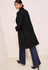 Palton lung negru cu un design structurat, cu închidere pe un singur rând de nasturi și buzunare laterale. Asortat cu blugi din denim albastru cu picioare largi și pantofi cu toc ascuțit negri.