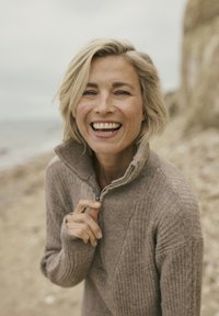 Femme blonde souriante tenant la fermeture éclair de son pull en laine marron sur une plage rocheuse avec une mer calme en arrière-plan.